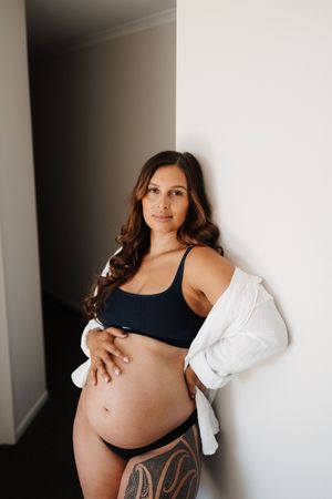 Expectant mother in black sports bra and white cardigan poses against wall showing pregnancy belly and leg tattoo.