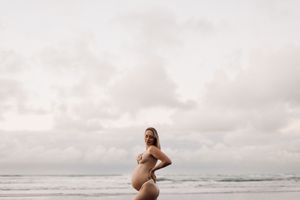 A silhouetted maternity figure stands on Raglan beach at sunset with cloudy skies in the background.