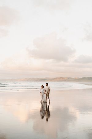 Two figures stand on reflective beach during pastel sunset with mountains in background.