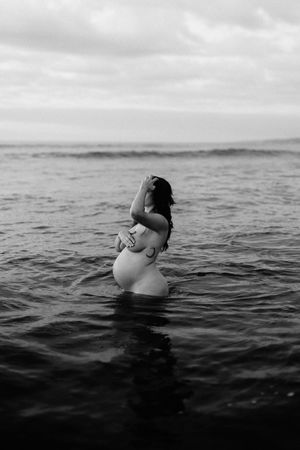 Black and white photo of pregnant woman standing in ocean water.