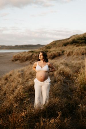 Expectant mother in white attire poses on coastal hillside during golden hour.