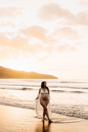 Pregnant woman in white swimwear poses on beach at sunset.