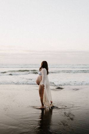 A silhouetted figure in a flowing dress stands at the ocean's edge during Raglan sunset.