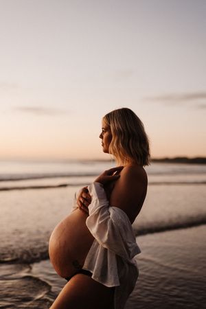Silhouetted pregnant woman in white dress stands at beach during golden hour.