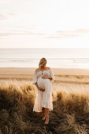 Pregnant woman in white dress poses among coastal dunes during sunset.