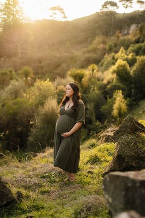 A pregnant woman in a green dress stands on a hillside during sunset.