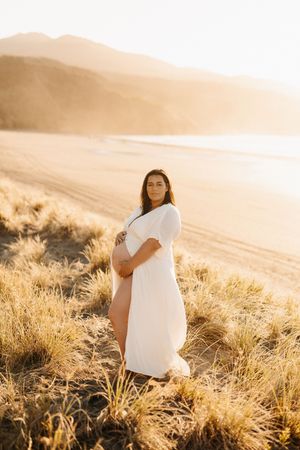 Maternity portrait of woman in white dress against golden coastal landscape.