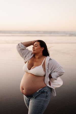 Pregnant person in white bra and denim jeans poses on misty beach at sunset.