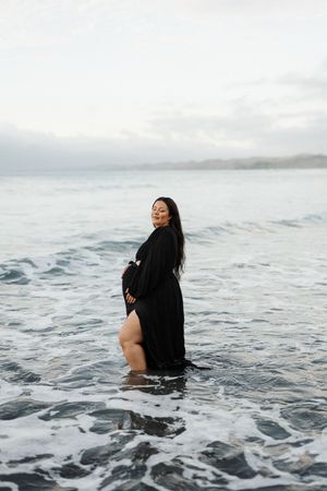 Woman in black dress standing in ocean waves during moody overcast day at the beach.