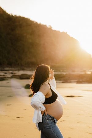 An expectant mother in casual attire poses on a beach during golden hour sunset.