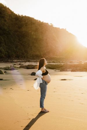 A pregnant woman in jeans and crop top stands on a beach at Raglan sunset.