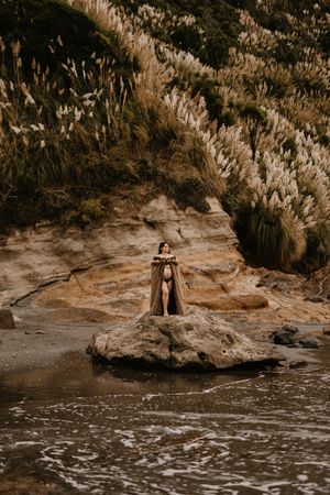 A person stands on a rocky outcrop surrounded by tall pampas grass near the ocean.