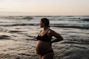 Silhouetted pregnant woman in black swimwear stands on beach at sunset with waves in background.