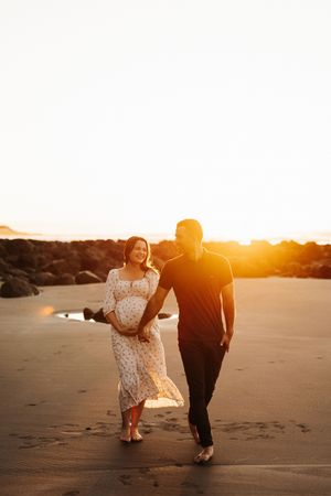Couple walking on beach at sunset during maternity photoshoot.