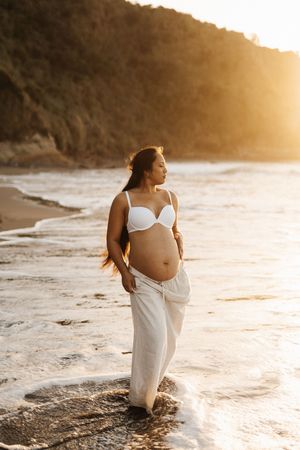 Pregnant woman in white attire poses on beach during golden sunset.