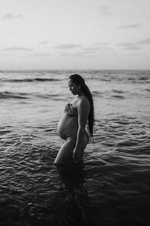 Black and white portrait of pregnant woman standing in ocean water.