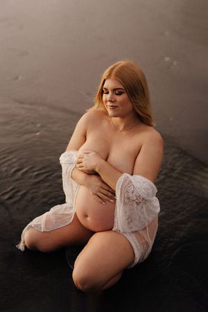 Expectant mother in white lace robe poses intimately on dark sandy beach.