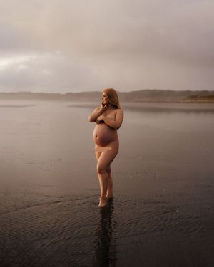 Silhouette of pregnant woman standing in shallow ocean water at dusk.