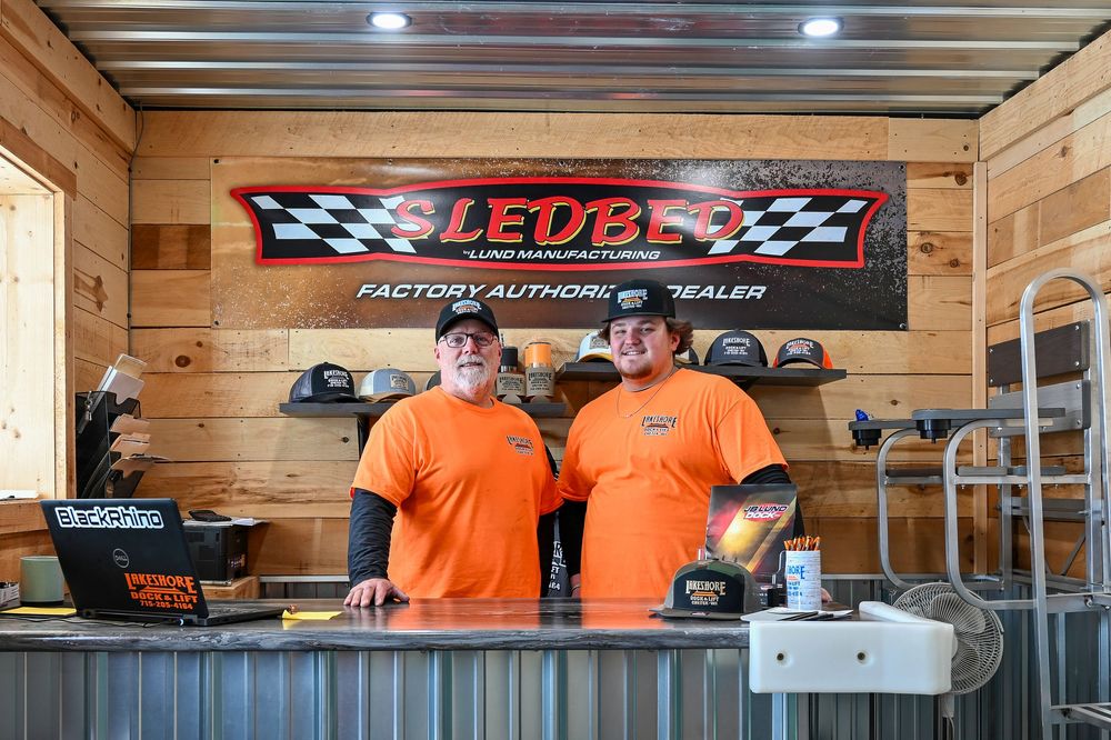 Two employees in orange shirts stand behind a counter at a Sledbed Lund Manufacturing authorized dealer showroom.
