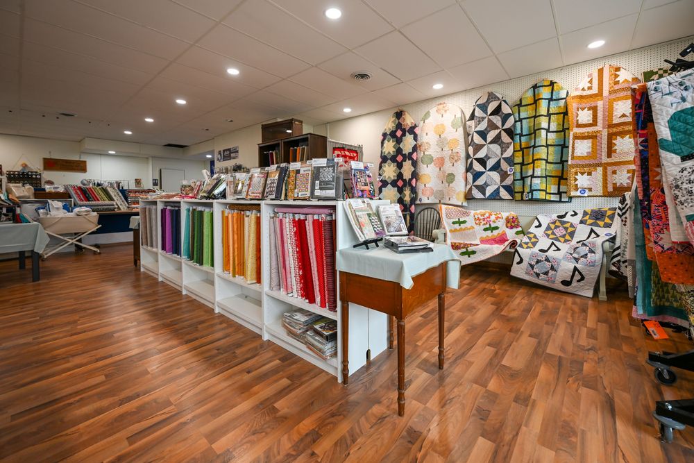 Colorful quilt fabric shop interior with organized shelves, vibrant fabric rolls, and quilts displayed on walls over hardwood floors.
