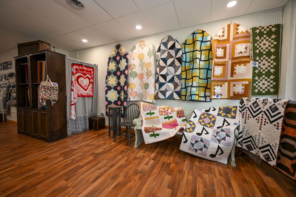 Colorful quilts displayed on walls and tables in a bright quilt shop with hardwood floors.
