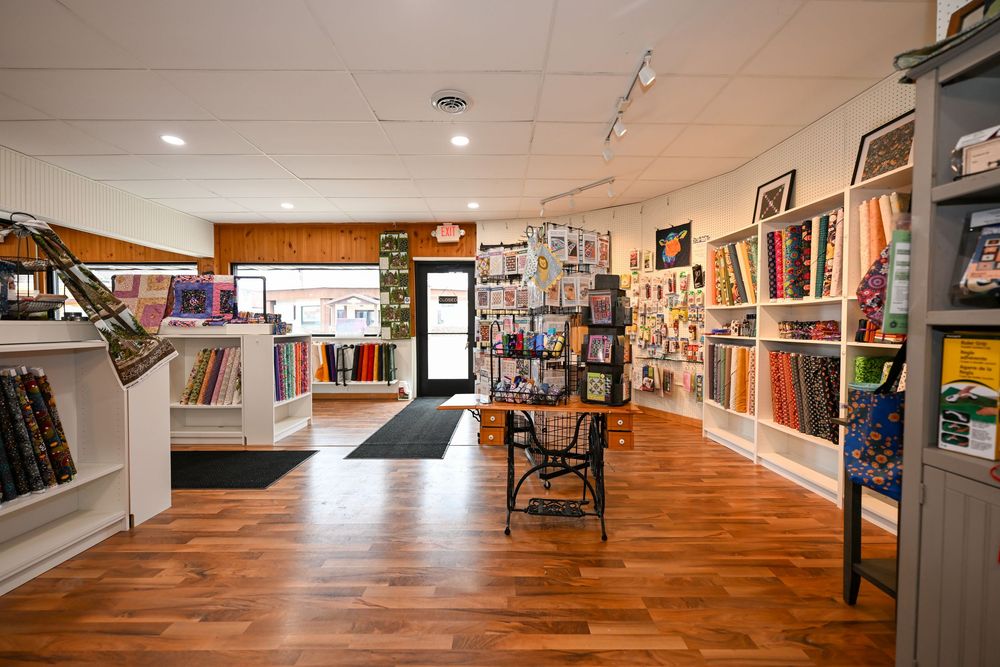 Bright fabric store interior with hardwood floors, colorful bolts of fabric on shelves, and display racks in a well-lit retail space.