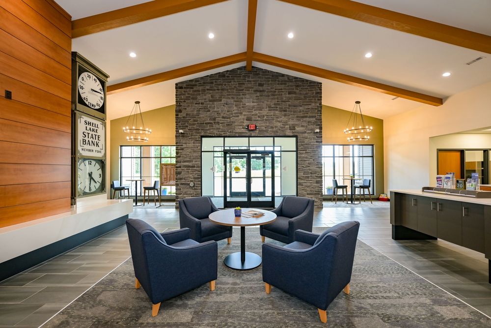 Modern hotel lobby with blue armchairs, stone accent wall, vaulted wood ceiling, and a round white table on a patterned rug.