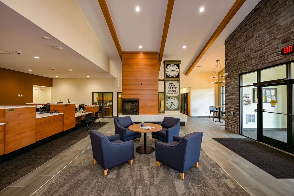 Modern hotel lobby with blue armchairs, round table, stone fireplace, wall clock, and reception desk on polished floors.