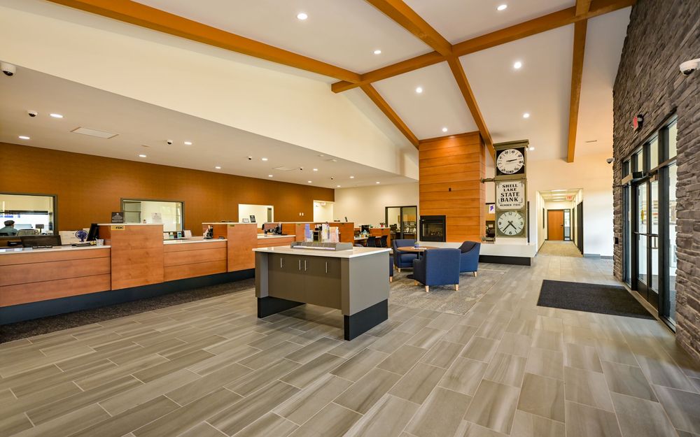 Modern bank lobby with wood beam ceilings, teller counters, hardwood floors, and a decorative clock near glass entrance doors.