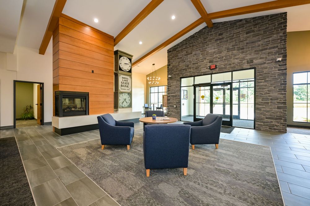 Modern hotel lobby with stone fireplace, large clock, blue armchairs, high ceilings, and floor-to-ceiling windows.