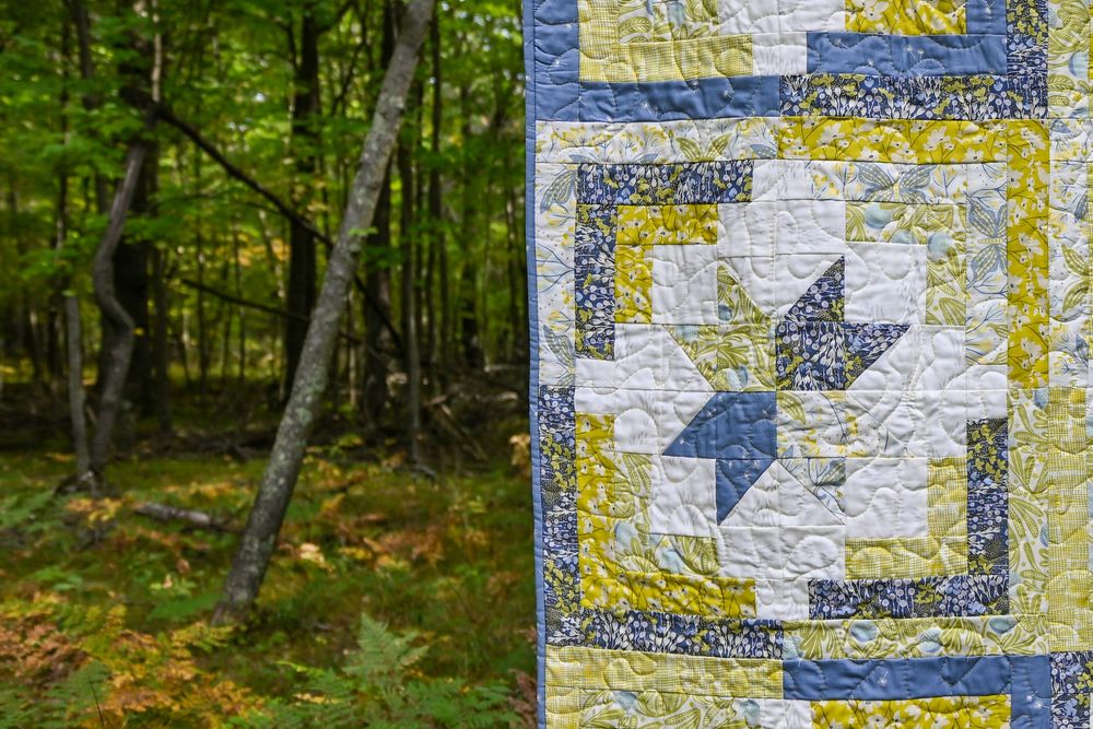 Blue and yellow patchwork quilt displayed outdoors against a lush green forest backdrop.