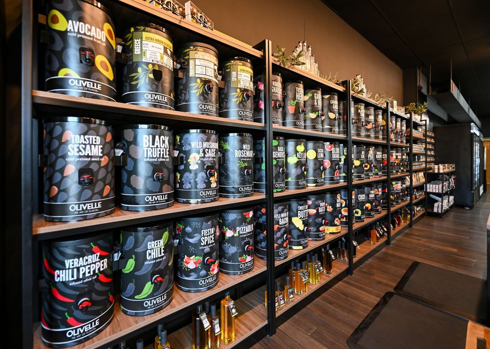 Shelves stocked with Olivelle branded olive oil tins and gourmet food products in a specialty grocery store.