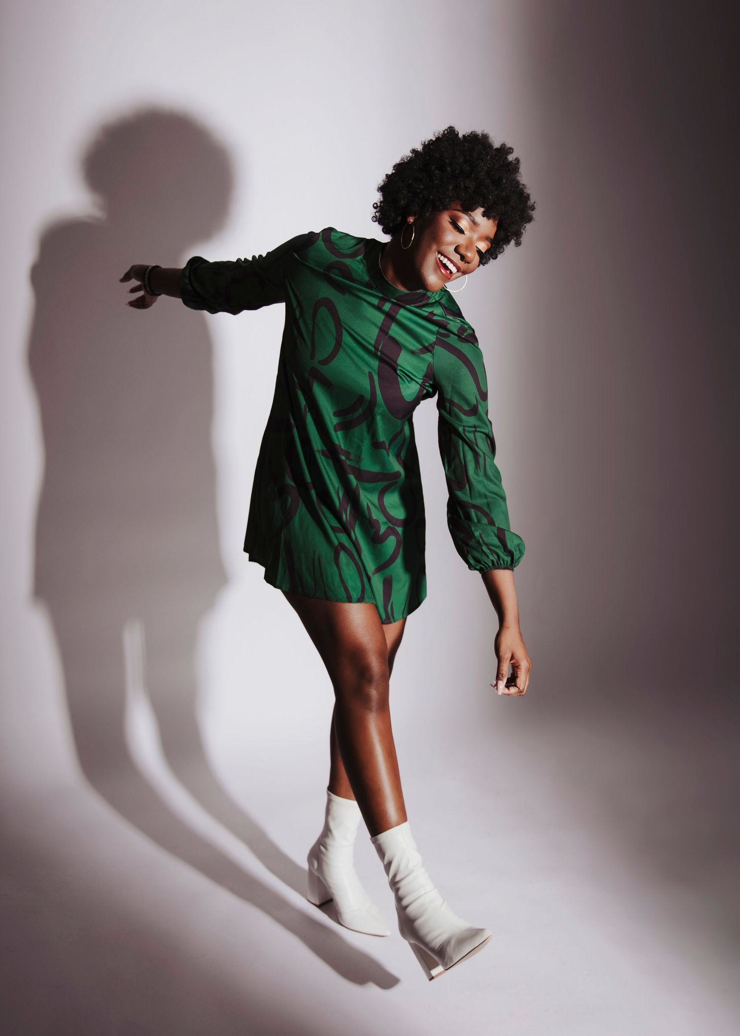 Fashion model in emerald green dress poses dynamically against wall creating dramatic shadow.