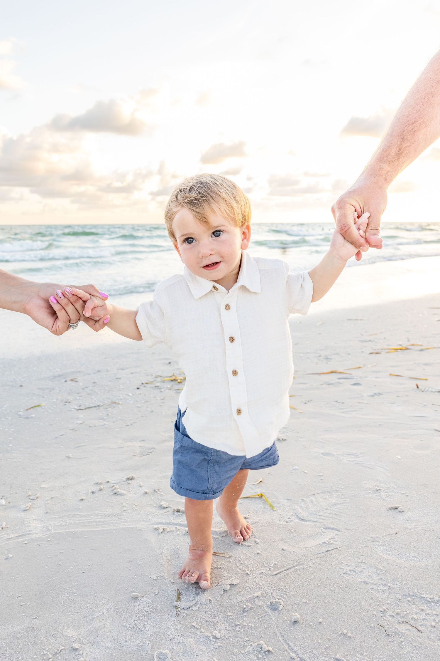 Adorable toddler in white shirt and blue shorts holds adults hands while standing on a sandy beach.