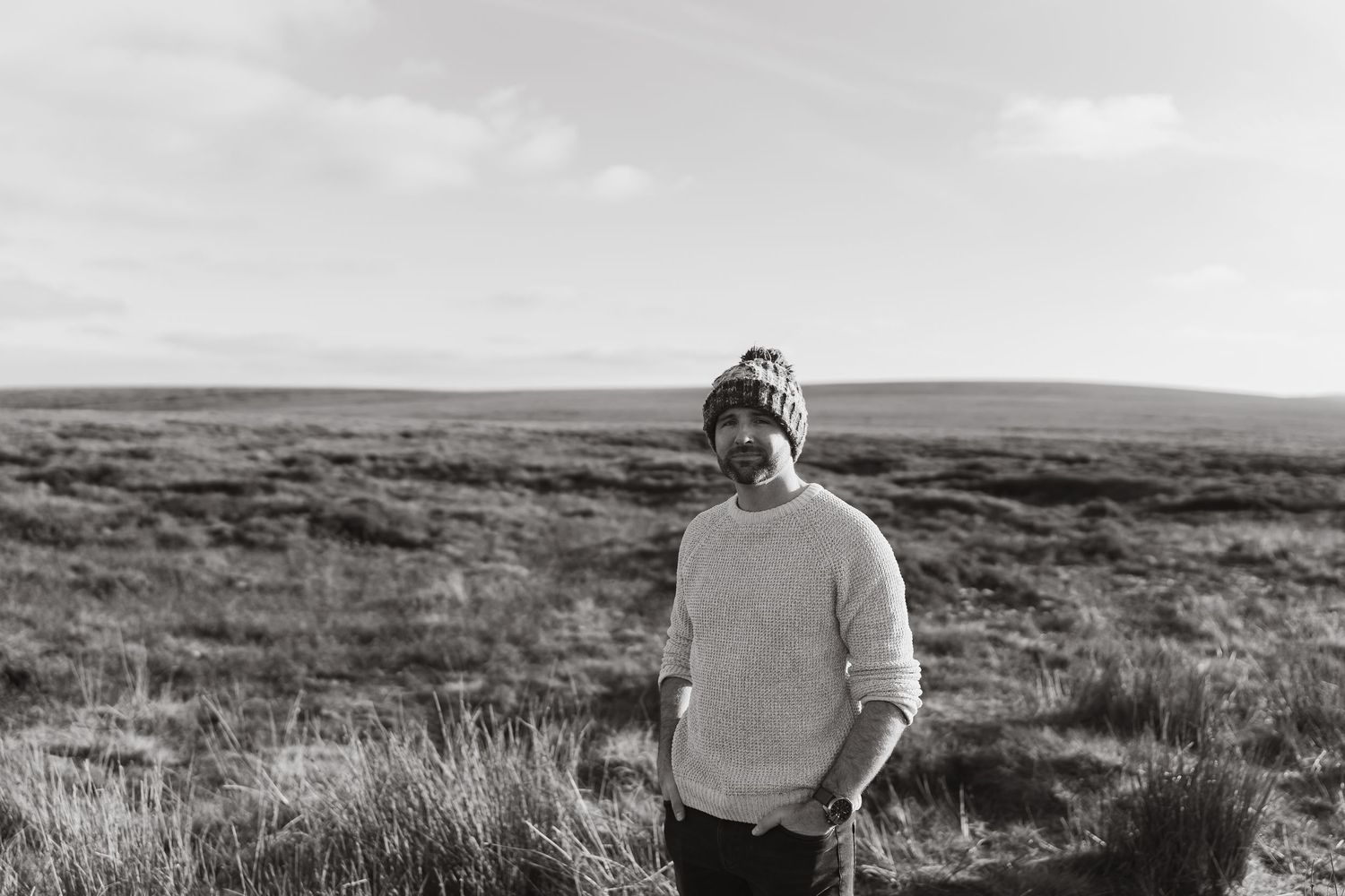 A person hikes through rolling hills covered in heather with a scenic moorland landscape stretching into the distance.