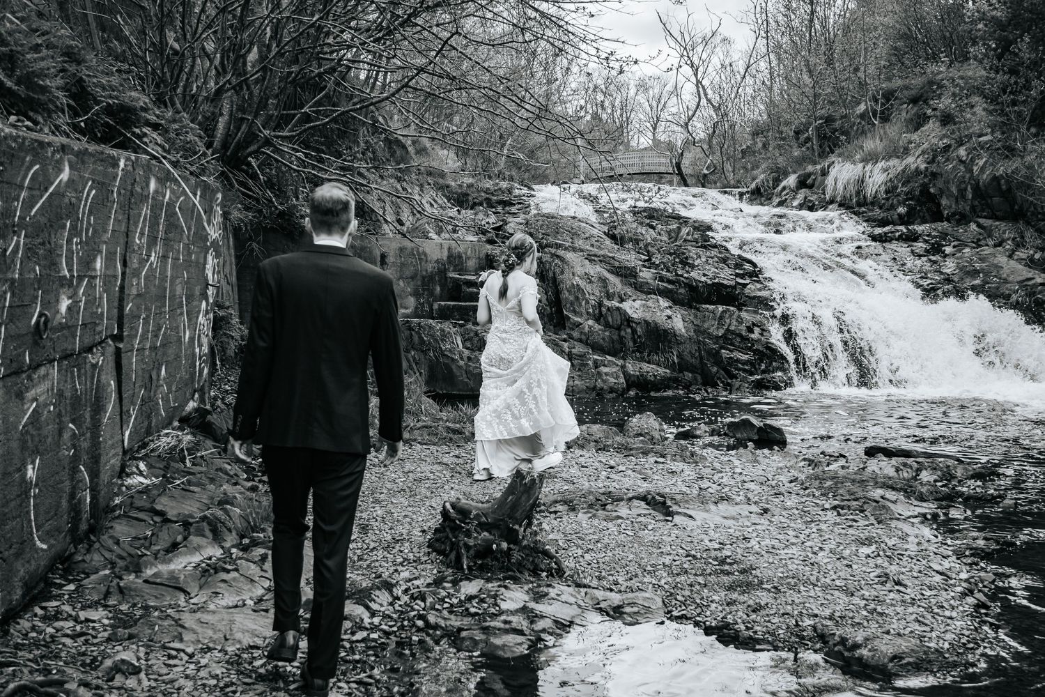 Black and white photograph of two figures standing near a waterfall on rocky terrain.