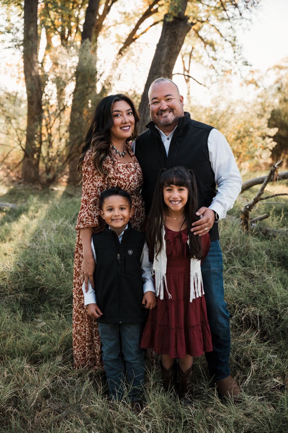 Family of five poses together in casual attire against a natural wooded backdrop during sunset.