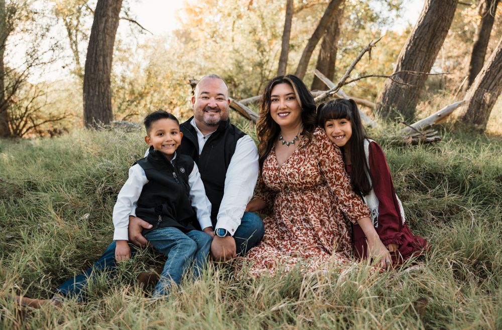 Family of four sits together on grassy hill during golden hour autumn photoshoot in wooded setting.