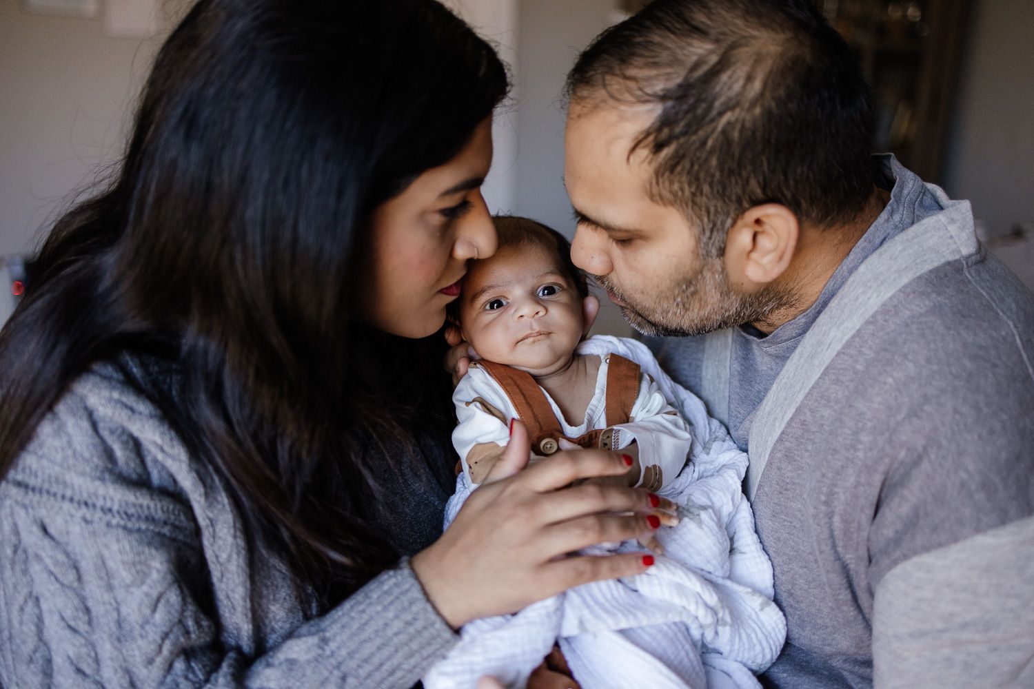 Parents kiss baby on his cheeks during their in-home lifestyle newborn photo shoot