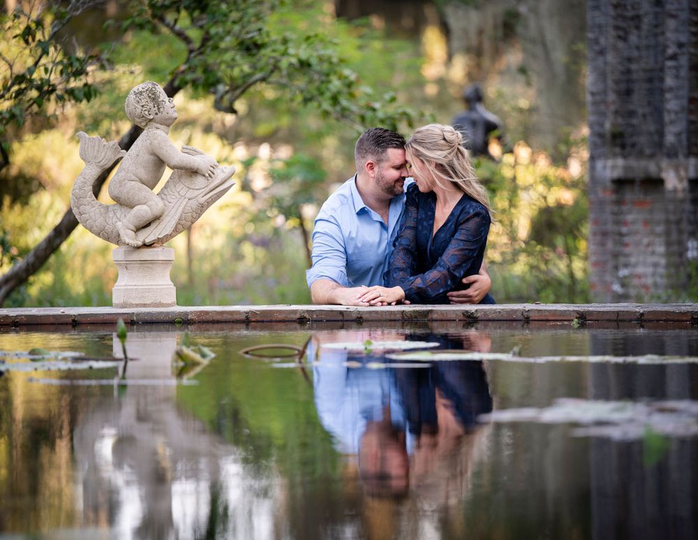 A couple shares an intimate moment by a reflective pond with a classical statue in the background.