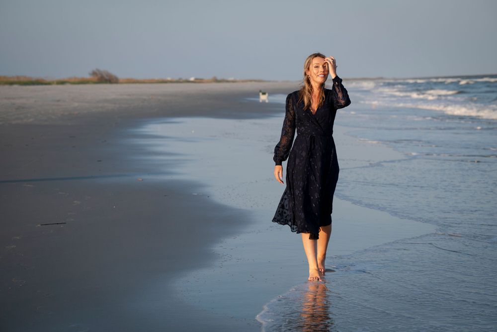 A woman in a flowing black dress walks along the shoreline at sunset on a peaceful beach.