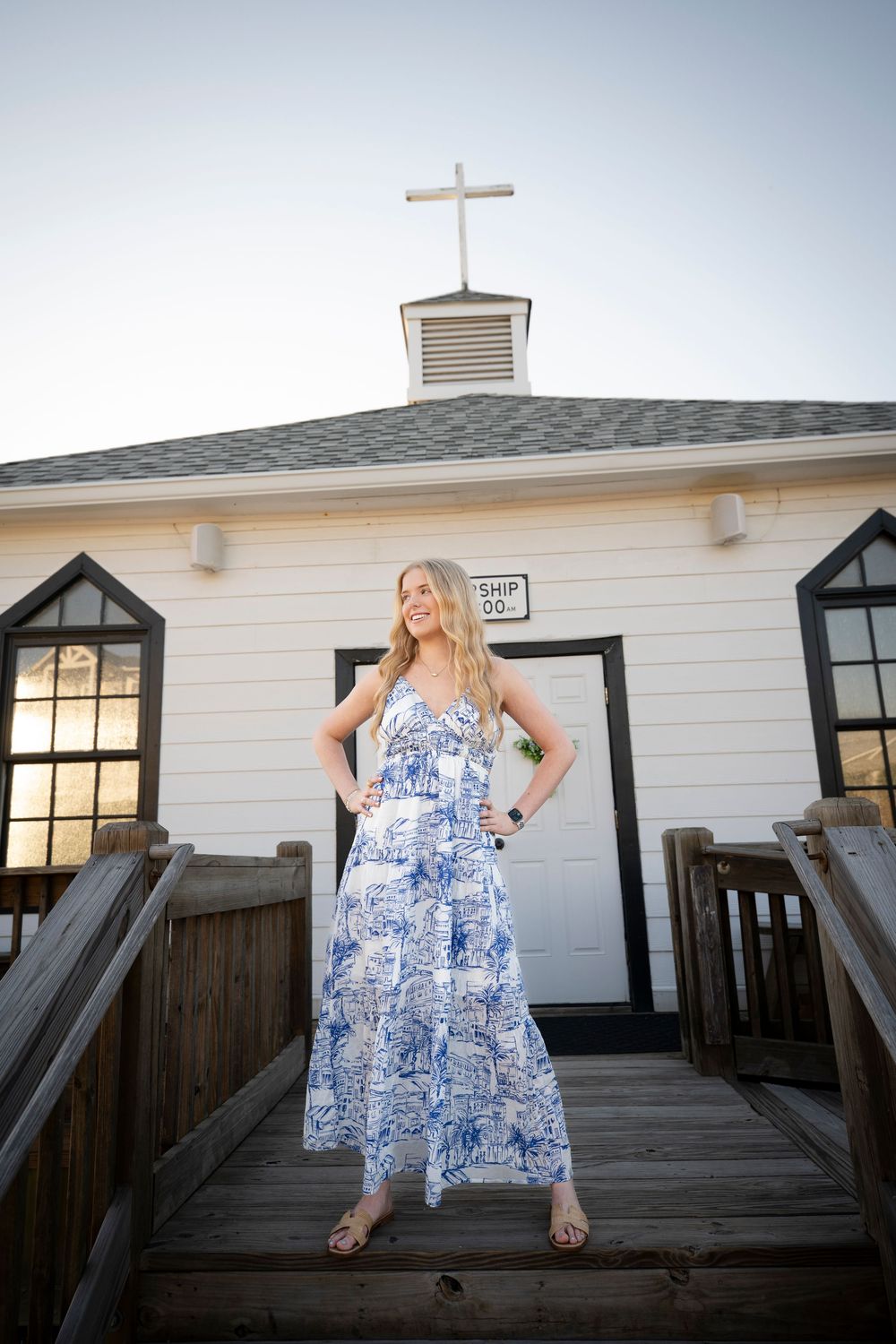 White chapel with cross on top and wooden deck platform at sunset with a figure in a blue and white floral maxi dress posing.