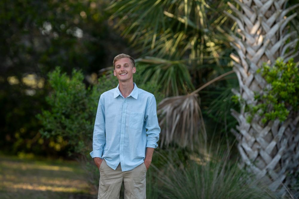 Young man in light blue shirt and khaki pants smiling outdoors among lush green palms and tropical foliage.