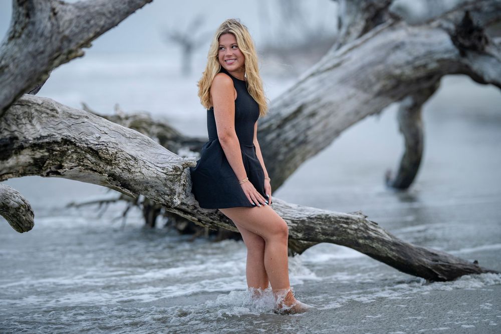 Blonde woman in black dress posing on driftwood at a misty beach shoreline.