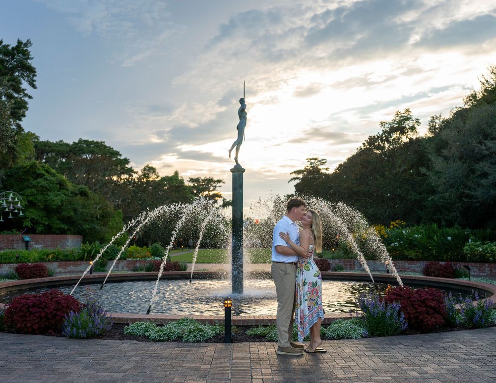 Couple sharing a romantic kiss in front of a fountain with a statue at sunset in a lush garden.