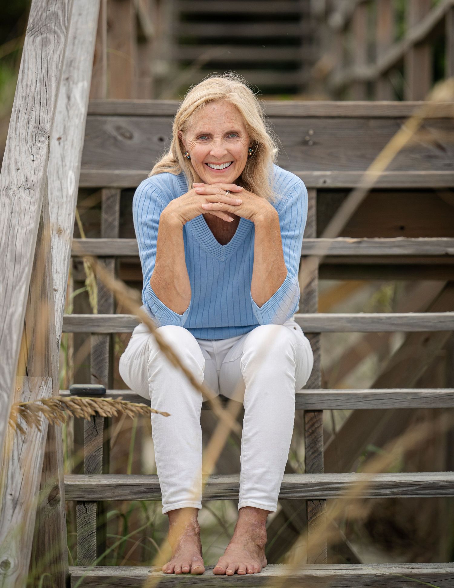 Smiling blonde woman in blue sweater and white pants sitting barefoot on wooden beach stairs.