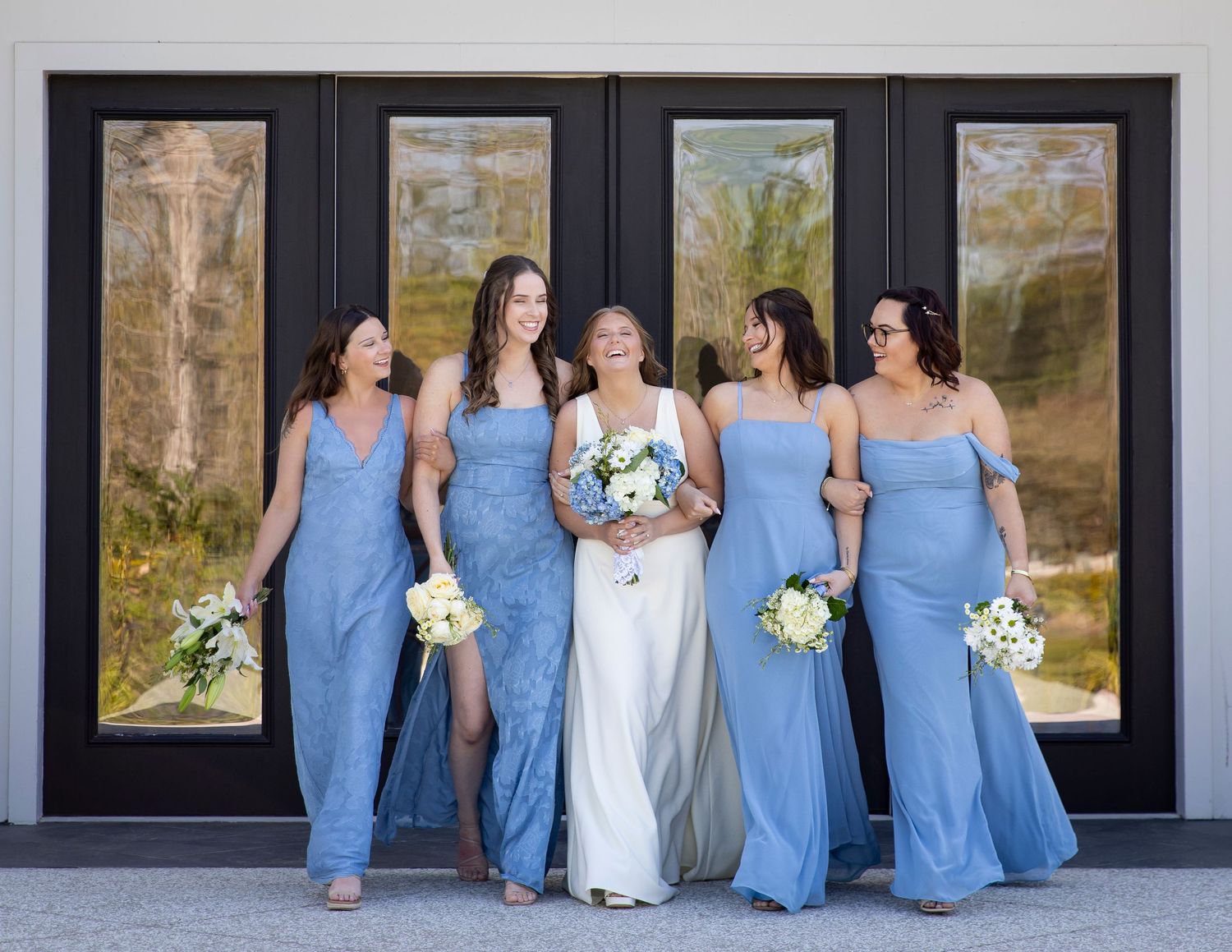 Bride in white gown with four bridesmaids in dusty blue dresses holding white bouquets posing outdoors.