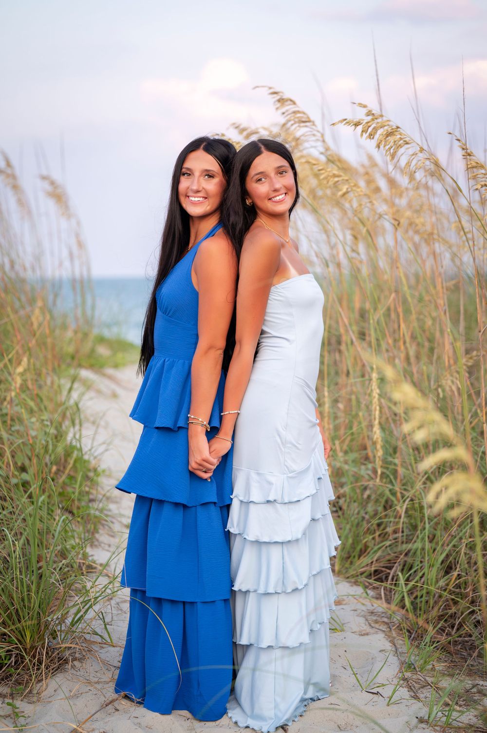 Two young women in blue and white tiered gowns pose together on a sandy beach path surrounded by sea oats at sunset.