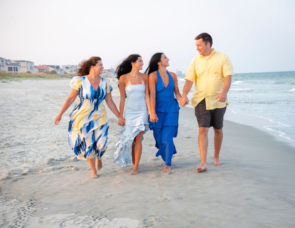 Four friends walking hand-in-hand on a sandy beach, wearing colorful summer dresses and casual attire.