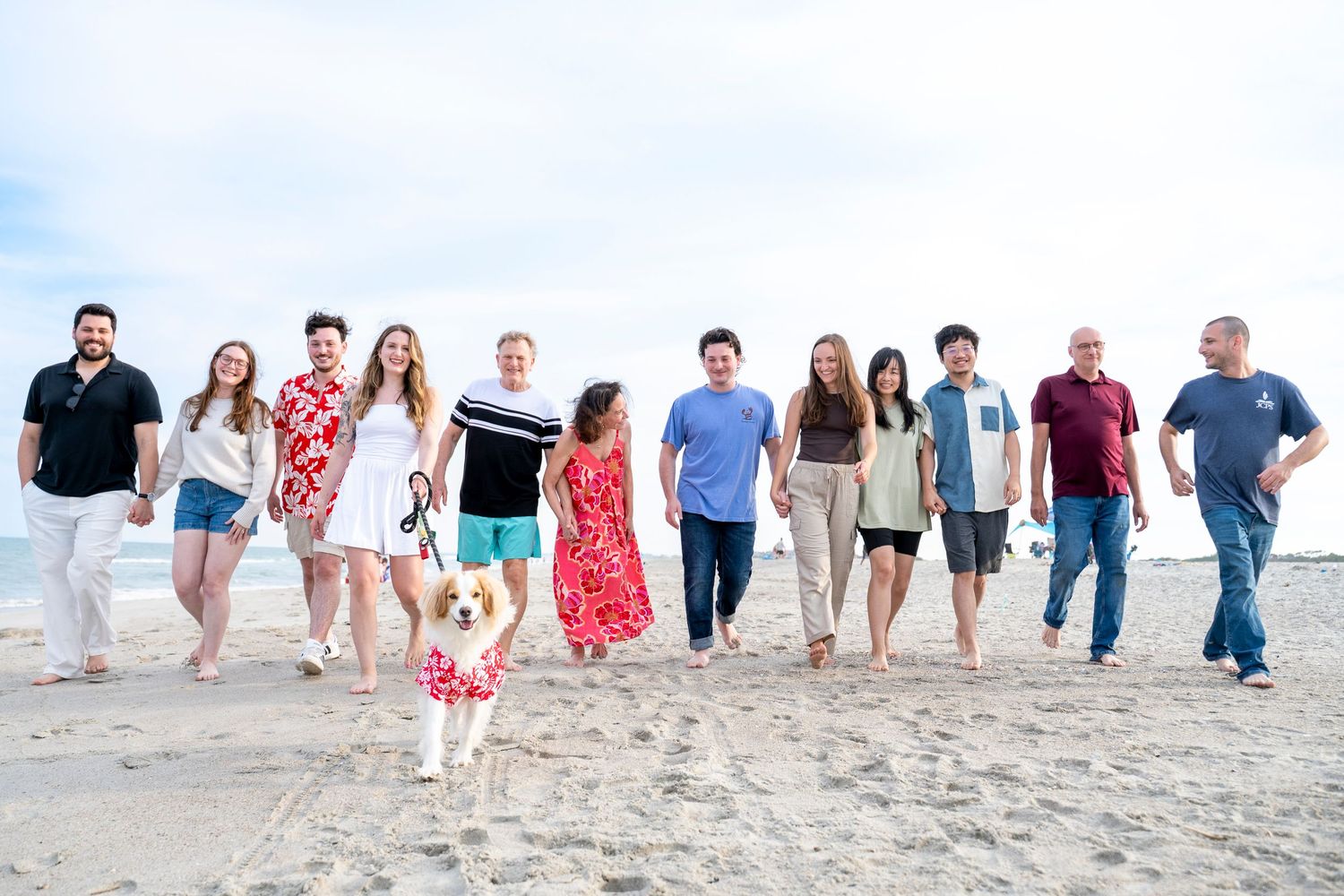 A large group of smiling people and a dog walk together on a sandy beach on a cloudy day.